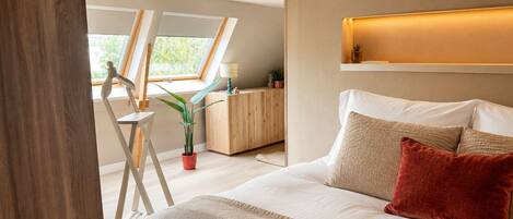 A bedroom with a bed, pillows, a bedside table, a lamp, a window, and a plant.