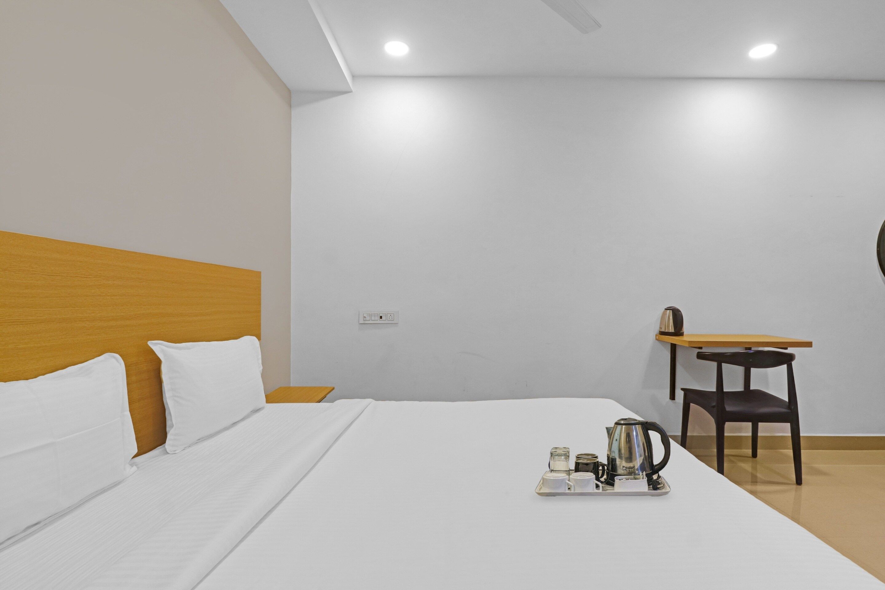 Habitación de hotel con cama, cabecero de madera, mesita con tetera y tazas, y una silla.