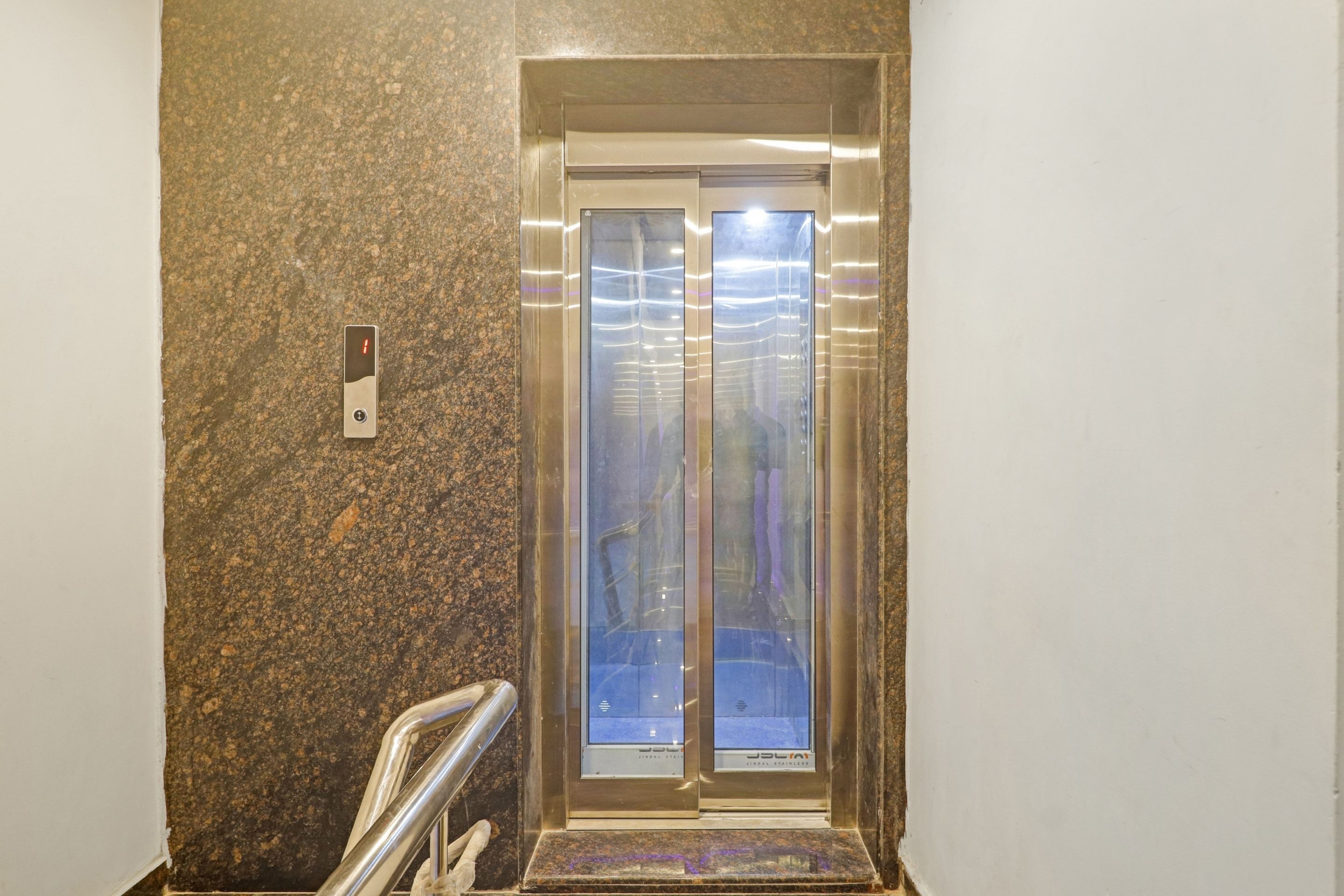 An elevator with a metallic finish and a glass door, situated in a hallway with a speckled floor.