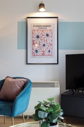 Een woonkamer met een lijstwerkprint van een bloemenmarkt, een blauwe fauteuil, een bruin kussen, een zwart tv-meubel en een potplant.