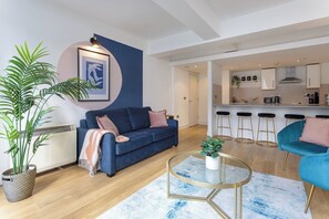 A modern living room with a blue sofa, a glass coffee table, and a kitchen area with bar stools. - Flatzy - Prime City Apartment 2 Baths Sleeps 6 (Merseyside)