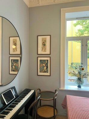 Ein Zimmer mit Klavier, gerahmten Kunstwerken an der Wand, einem Sessel und einem Fenster mit Blick auf Grünflächen.