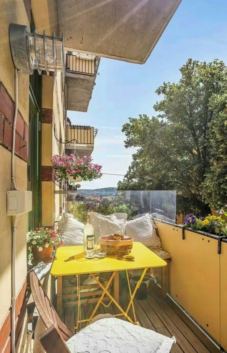 Een klein balkon met een gele tafel, een fles en een schaal. Er staan potplanten en er is uitzicht op een boom en gebouwen.