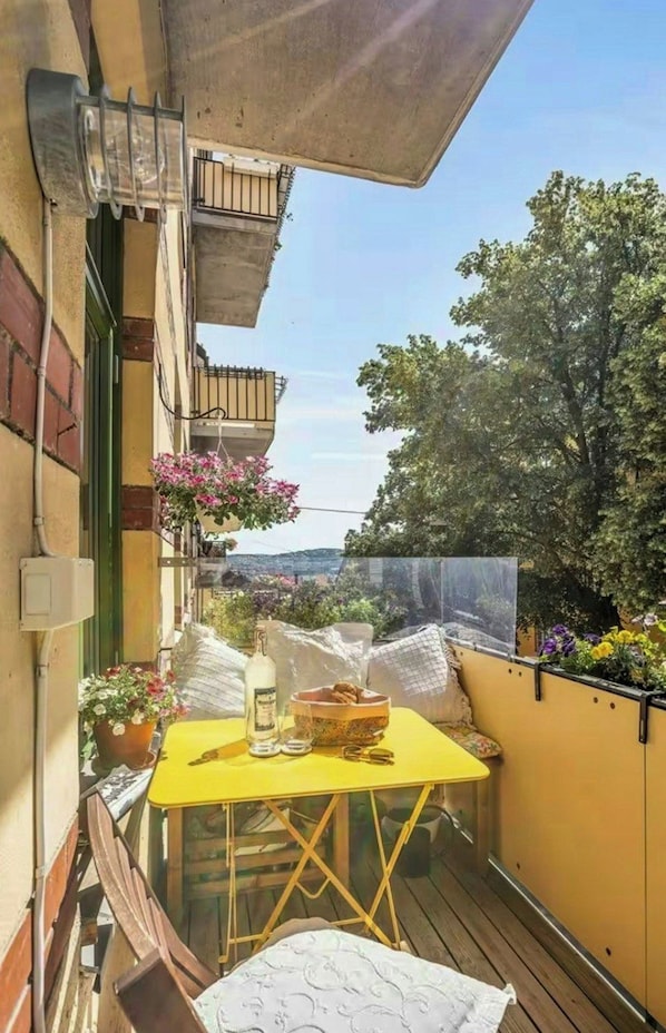 Ein kleiner Balkon mit einem gelben Tisch, einer Flasche und einer Schüssel. Es gibt Blumentöpfe und einen Blick auf einen Baum und Gebäude.