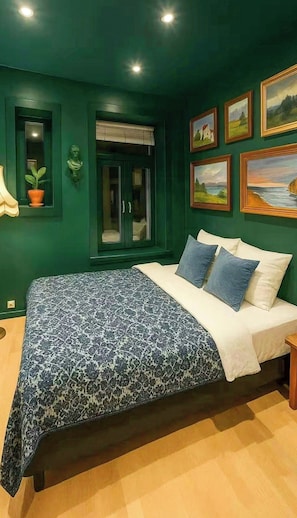 Ein Schlafzimmer mit einem Bett, einem Fenster mit Jalousien, einer Zimmerpflanze und gerahmten Bildern an der Wand.