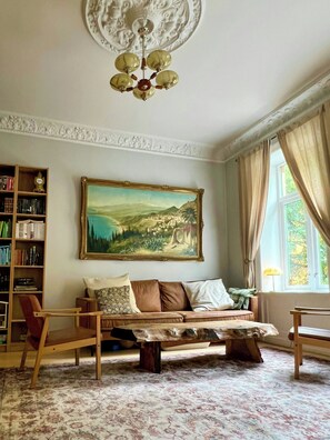 Ein Wohnzimmer mit einer Couch, Sesseln, einem Couchtisch, einem Bücherregal, einem Gemälde und einem Fenster mit Vorhängen.
