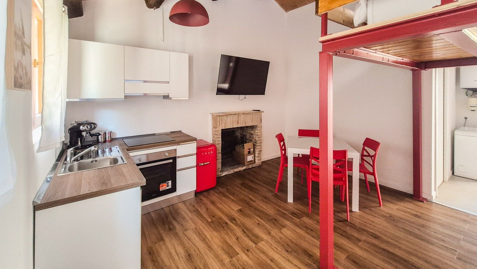 A compact kitchen with a built-in oven, a red refrigerator, and a dining area with red chairs.