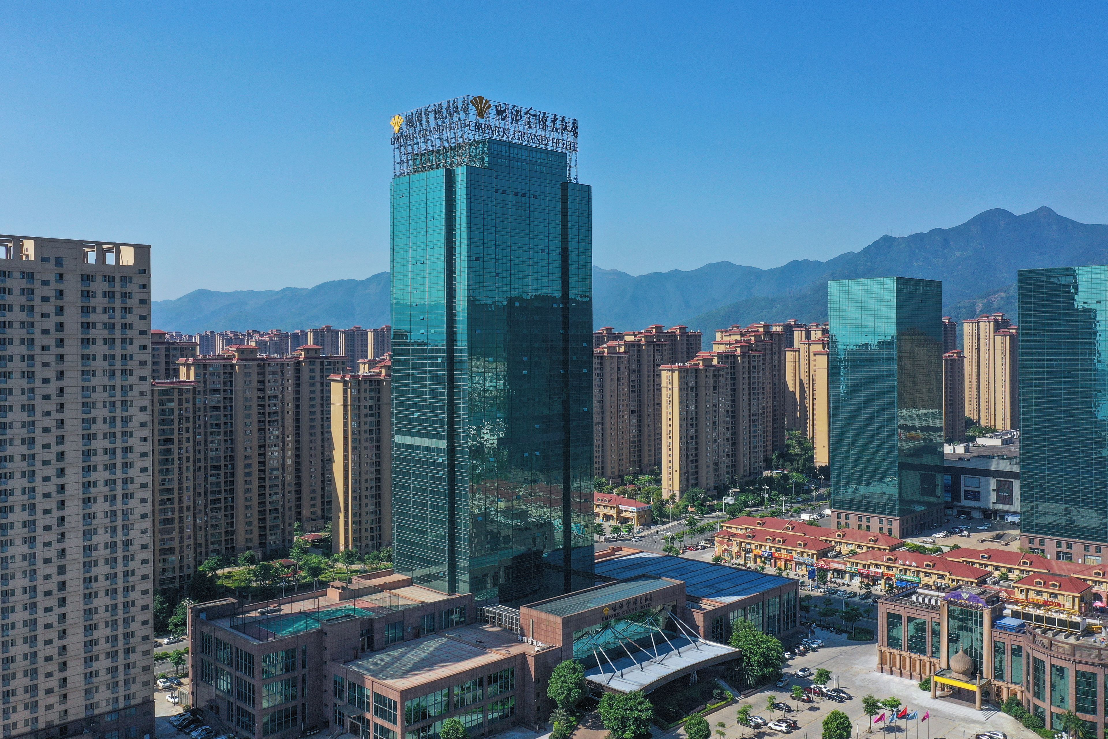 A high-rise hotel with a modern glass facade, surrounded by other high-rise buildings, set against a mountainous backdrop.