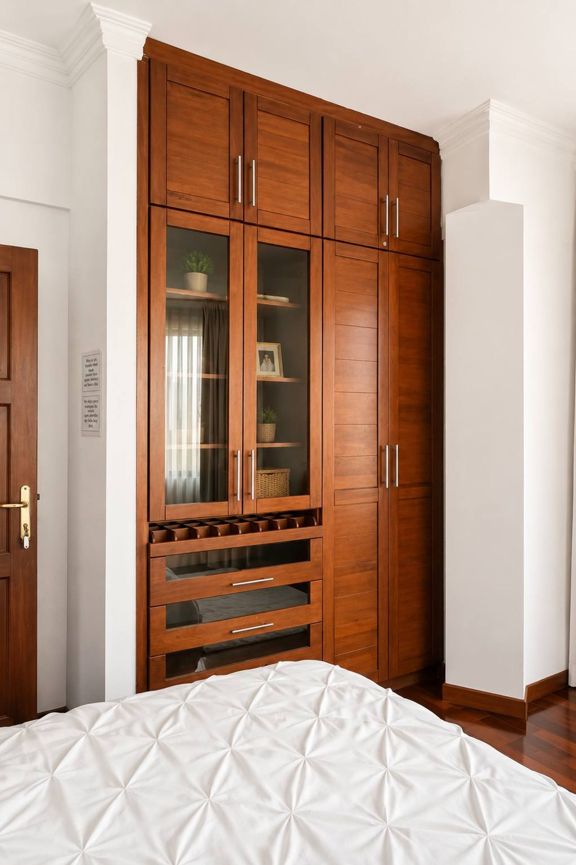 A bedroom with a large wooden wardrobe, a bed with a white quilt, and a small window with curtains.