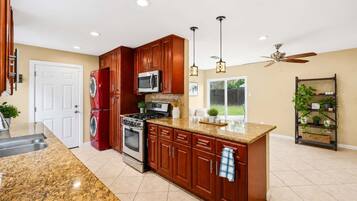 A kitchen with granite countertops, wooden cabinets, stainless steel appliances, pendant lighting, and a ceiling fan.