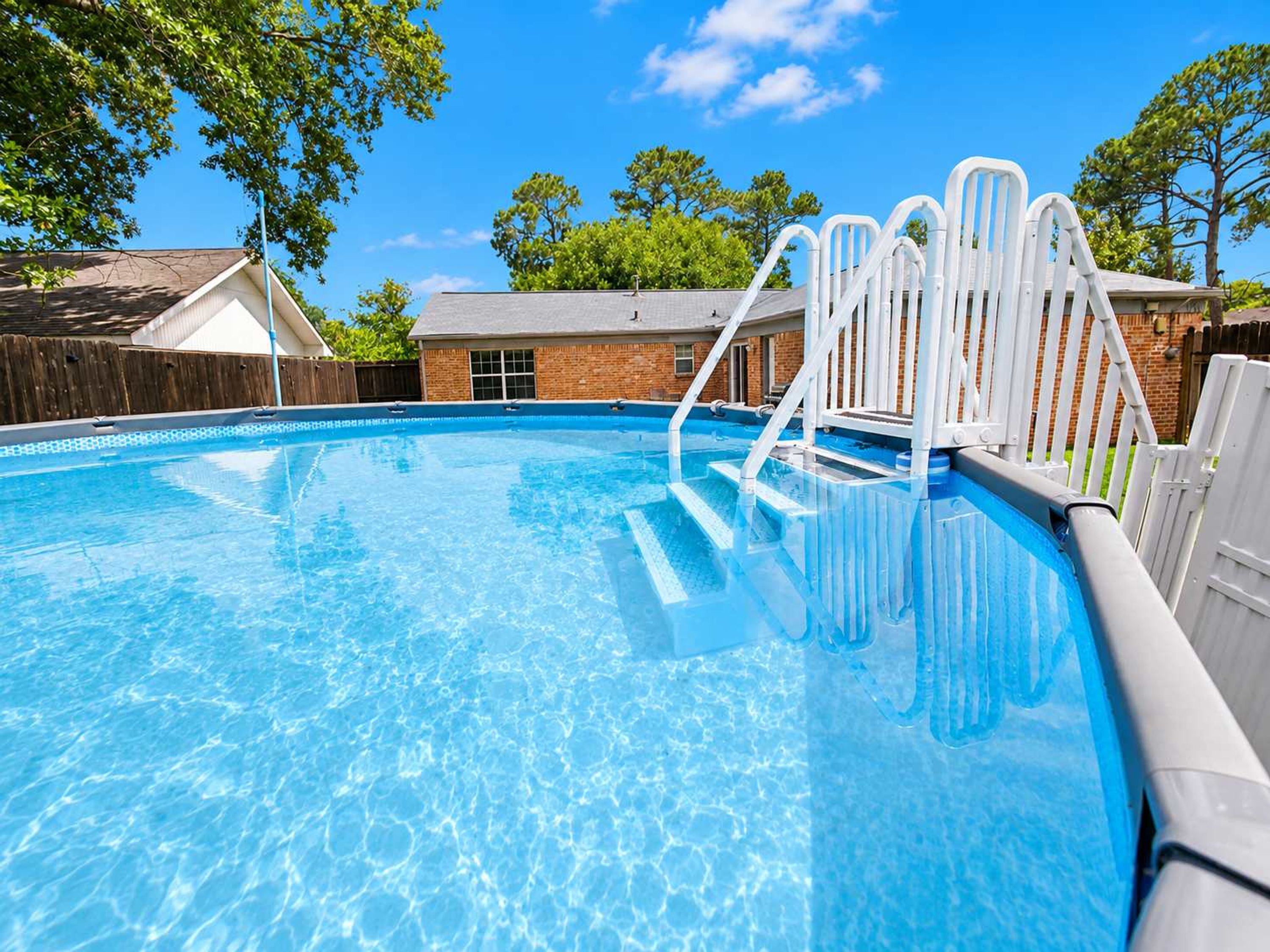 A clear swimming pool with steps leading into it, surrounded by a wooden fence and trees.
