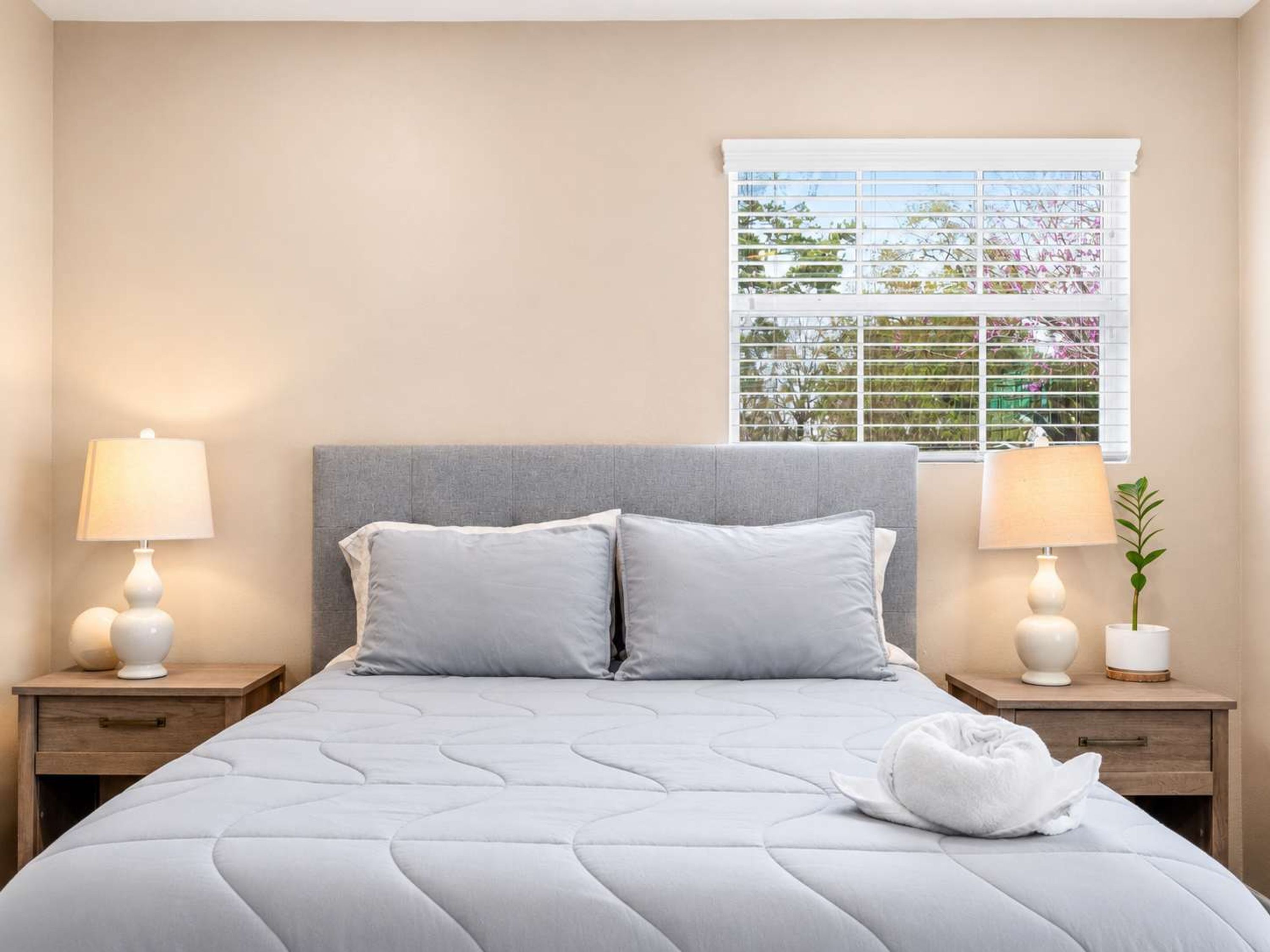 A hotel room with a bed, two bedside tables with lamps, a window with blinds, and a view of greenery outside.