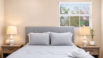 A hotel room with a bed, two bedside tables with lamps, a window with blinds, and a view of greenery outside.