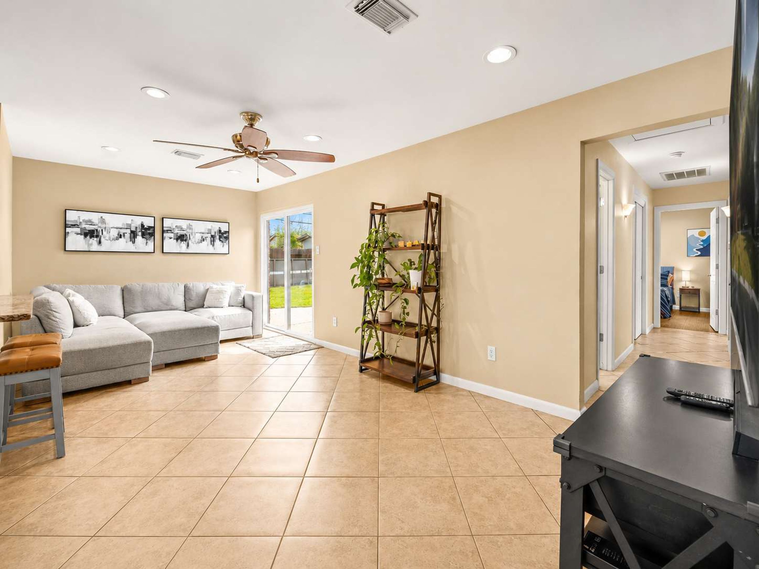 A spacious living room with a grey sofa, a ceiling fan, a wooden plant stand, and a large window.