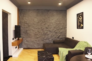 A modern living room with a stone-textured wall, a flat-screen TV, a brown sofa, a green throw, and a glass of wine on a table. - Cozy 1-bedroom apartment in charming Yerevan with AC (Yerevan)