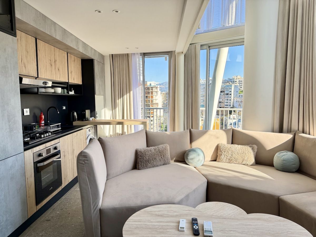 A modern living room with a sofa and a coffee table, a kitchen with wooden cabinets, and a city view through large windows.
