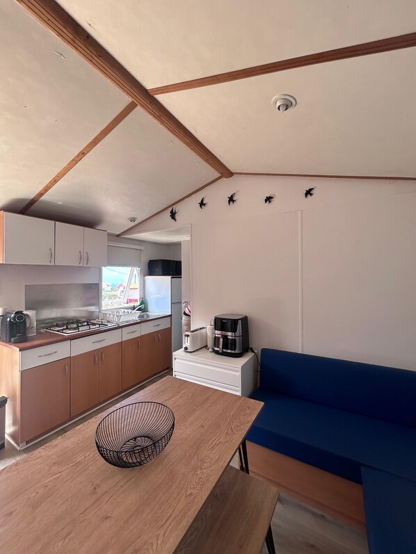 A compact kitchen with a stove, toaster, and coffee maker, a dining table with a wire basket, and a blue sofa.