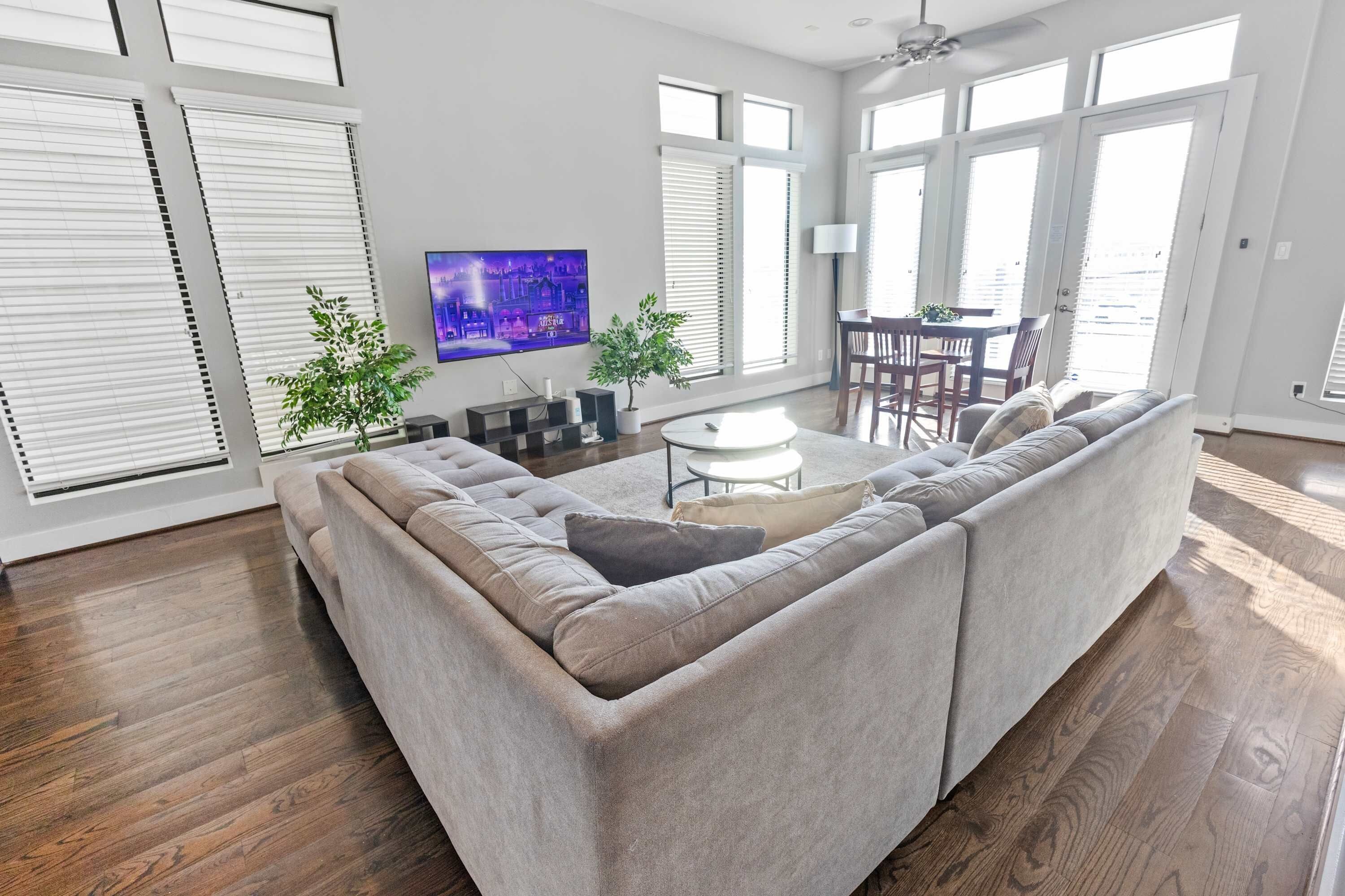A modern living room with a sectional sofa, a flat-screen TV, a dining area with a table and chairs, and large windows with blinds.