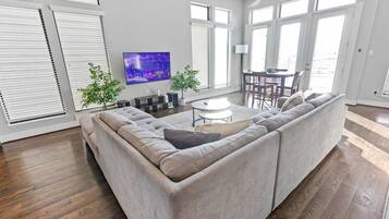 A modern living room with a sectional sofa, a flat-screen TV, a dining area with a table and chairs, and large windows with blinds.
