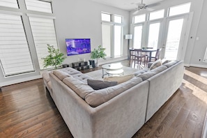 Uma sala de estar moderna com um sofá modular, uma TV de tela plana, uma área de jantar com mesa e cadeiras, e janelas grandes com persianas.