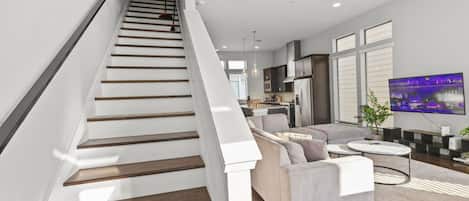 A modern living room with a staircase, a flat-screen TV, and a kitchen area.