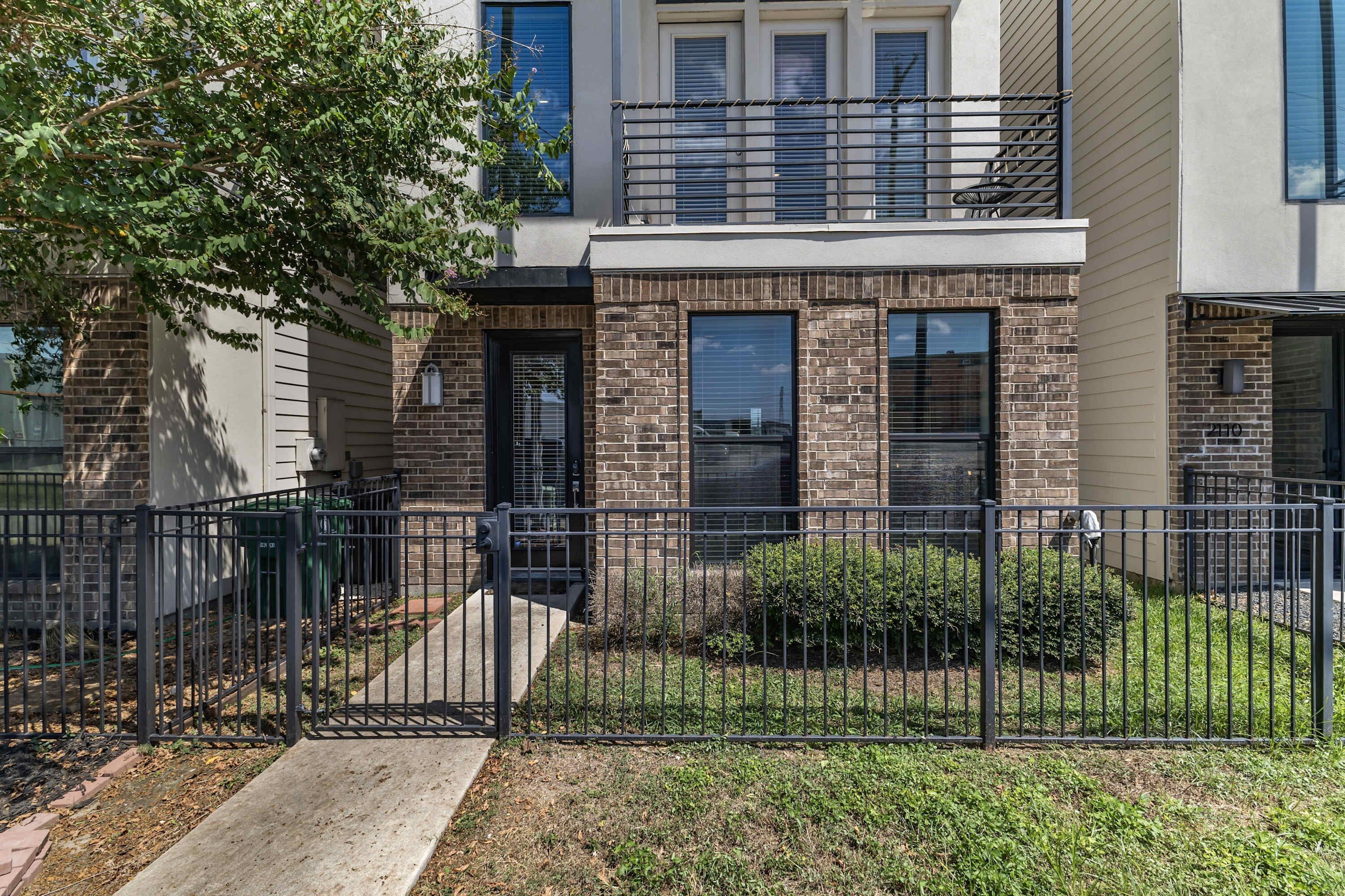A two-story residential building with a brick facade, a black metal fence, and a small front yard with grass and shrubs.