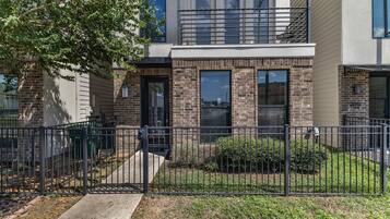 A two-story residential building with a brick facade, a black metal fence, and a small front yard with grass and shrubs.