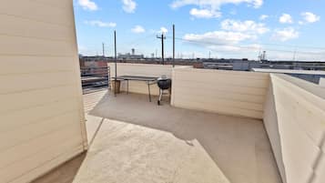 A rooftop patio with a barbecue grill and a table, surrounded by white walls and a clear sky.