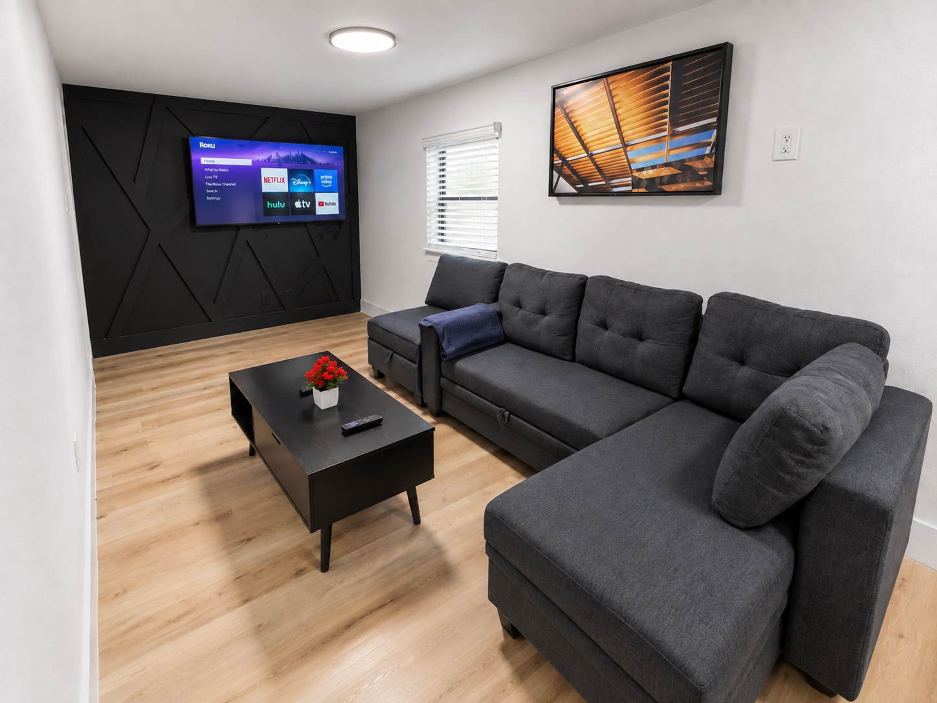 Una sala moderna con un sofá modular, una mesa de centro con un florero, un televisor de pantalla plana montado en una pared negra y un cuadro sobre el sofá.