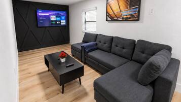 A modern living room with a sectional sofa, a coffee table with a vase, a flat-screen TV mounted on a black wall, and a painting above the sofa.