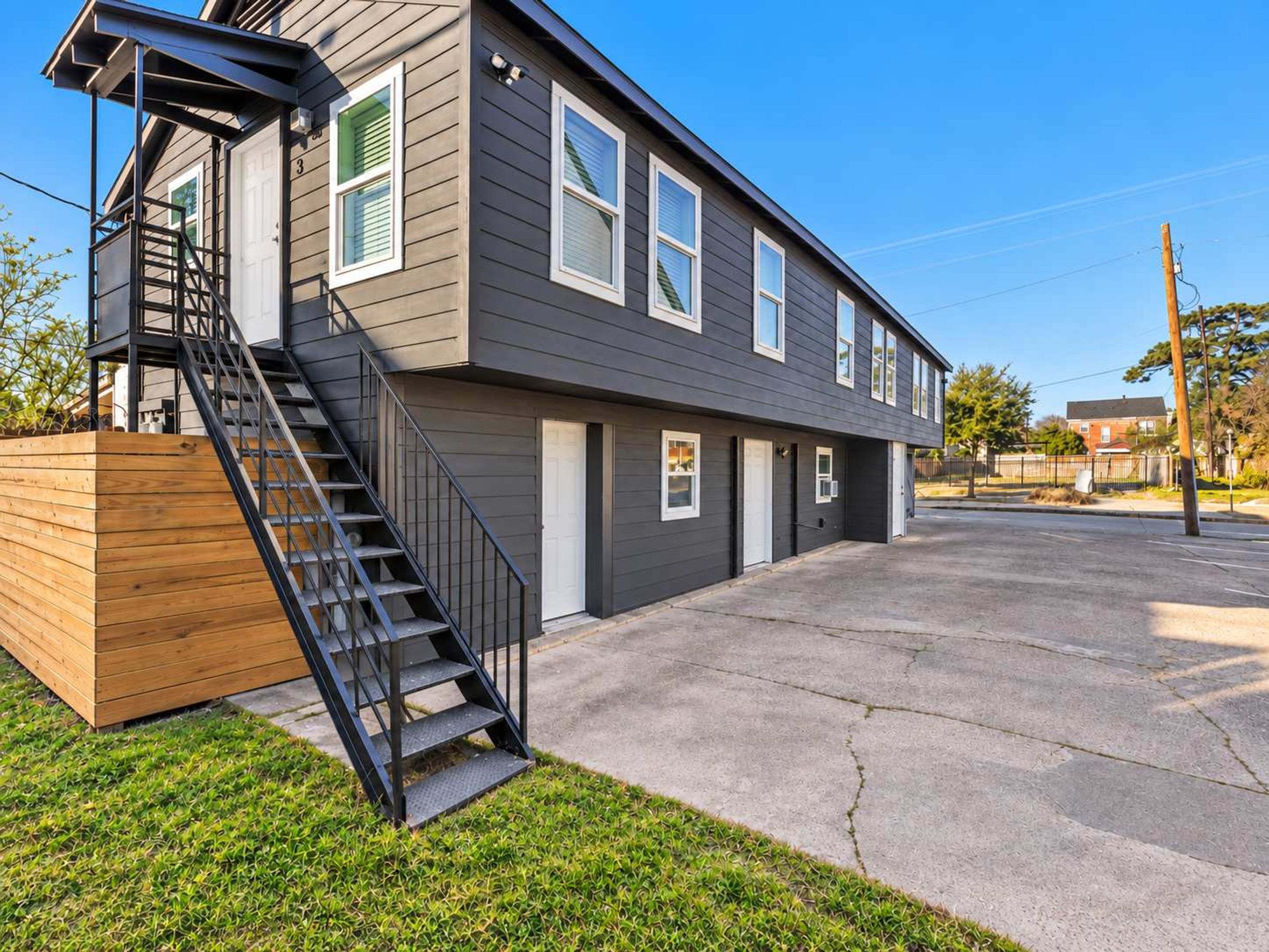 A two-story residential building with a wooden deck, a concrete driveway, and a metal staircase.