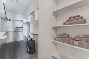 Quarto de hotel com carpete cinza, um saco de roupa suja pendurado, toalha branca e uma estante com toalhas dobradas.