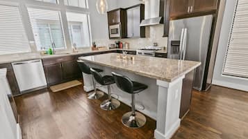 A modern kitchen with a central island, bar stools, stainless steel appliances, and a window with blinds.