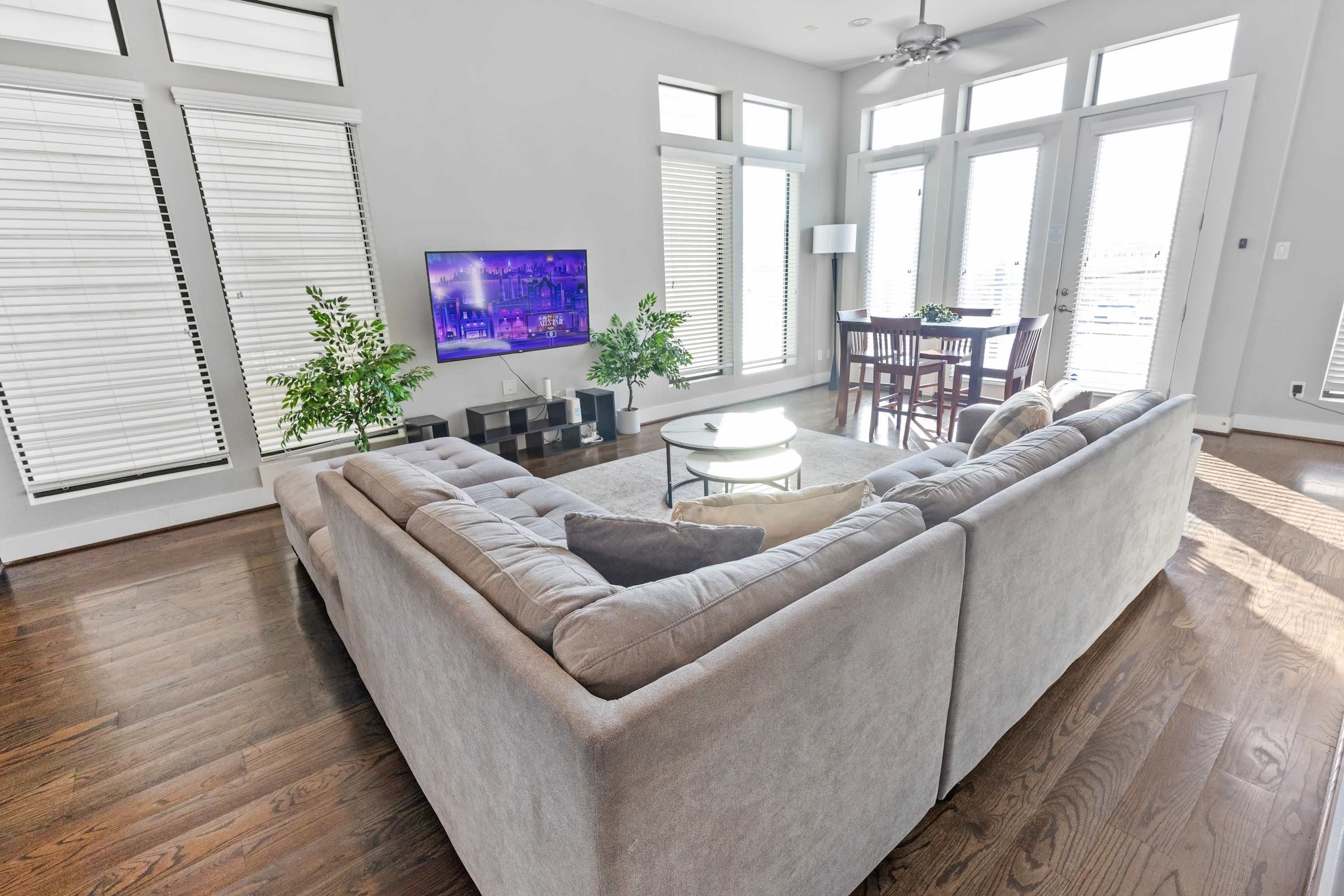 A modern living room with a sectional sofa, a flat-screen TV, a dining area with a table and chairs, and large windows with blinds.