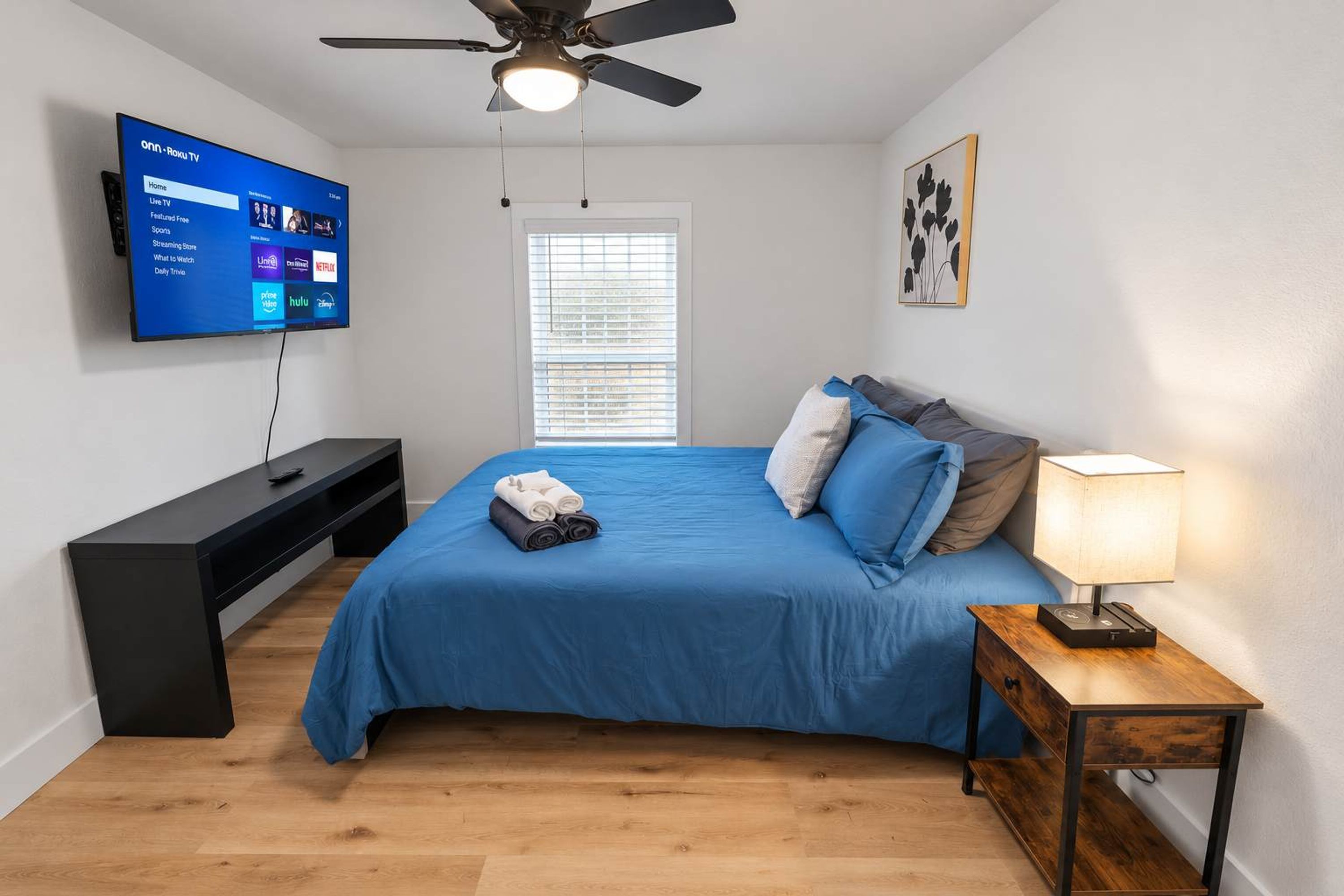 Un dormitorio con cama, televisor montado en la pared, ventilador de techo, mesita de noche con lámpara y ventana con persianas.