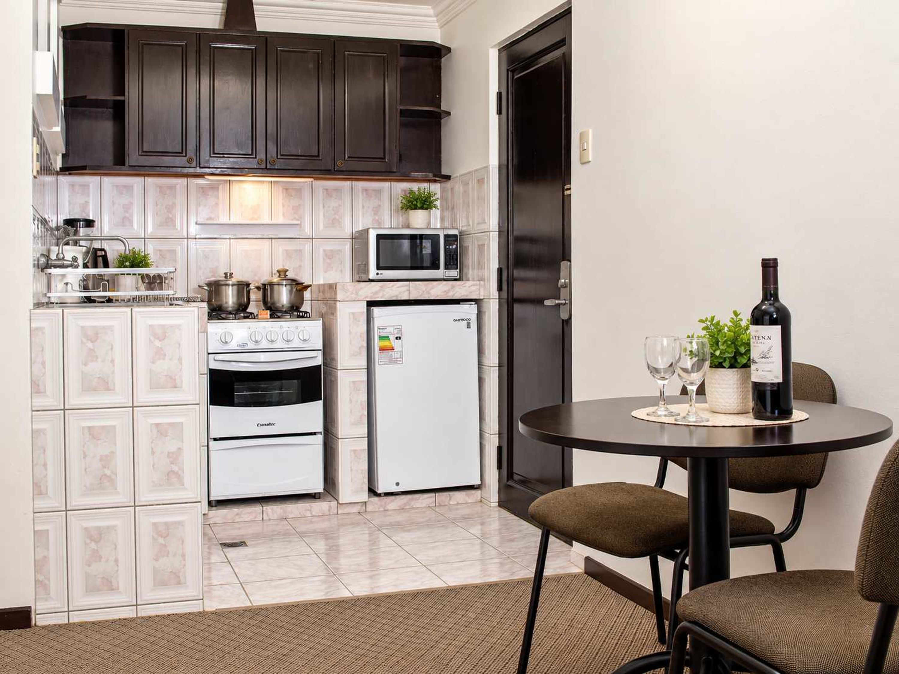 Une cuisine compacte équipée d’une cuisinière, d’un four, d’un four à micro-ondes et d’un réfrigérateur. On y trouve une table à manger avec des chaises et une bouteille de vin.