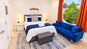 Een moderne slaapkamer met een groot bed, een blauwe bank, een kleine tafel en een raam met gordijnen.