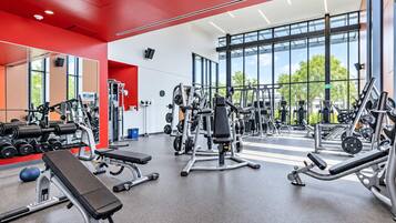 A modern gym with various exercise equipment, large windows, and a red accent wall.