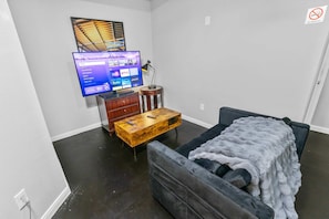 Ein kleiner, möblierter Raum mit einem an der Wand befestigten Fernseher, einem Holztisch, einer Couch und einem kleinen Beistelltisch.