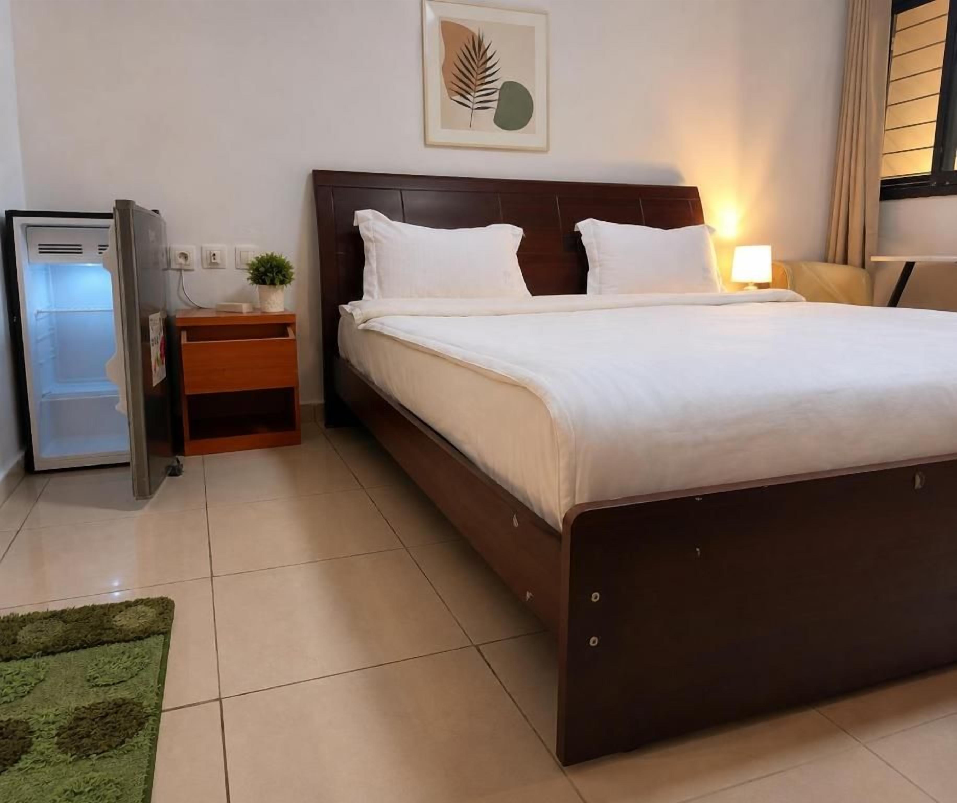 A hotel room with a bed, a nightstand, a lamp, a refrigerator, and a green rug.
