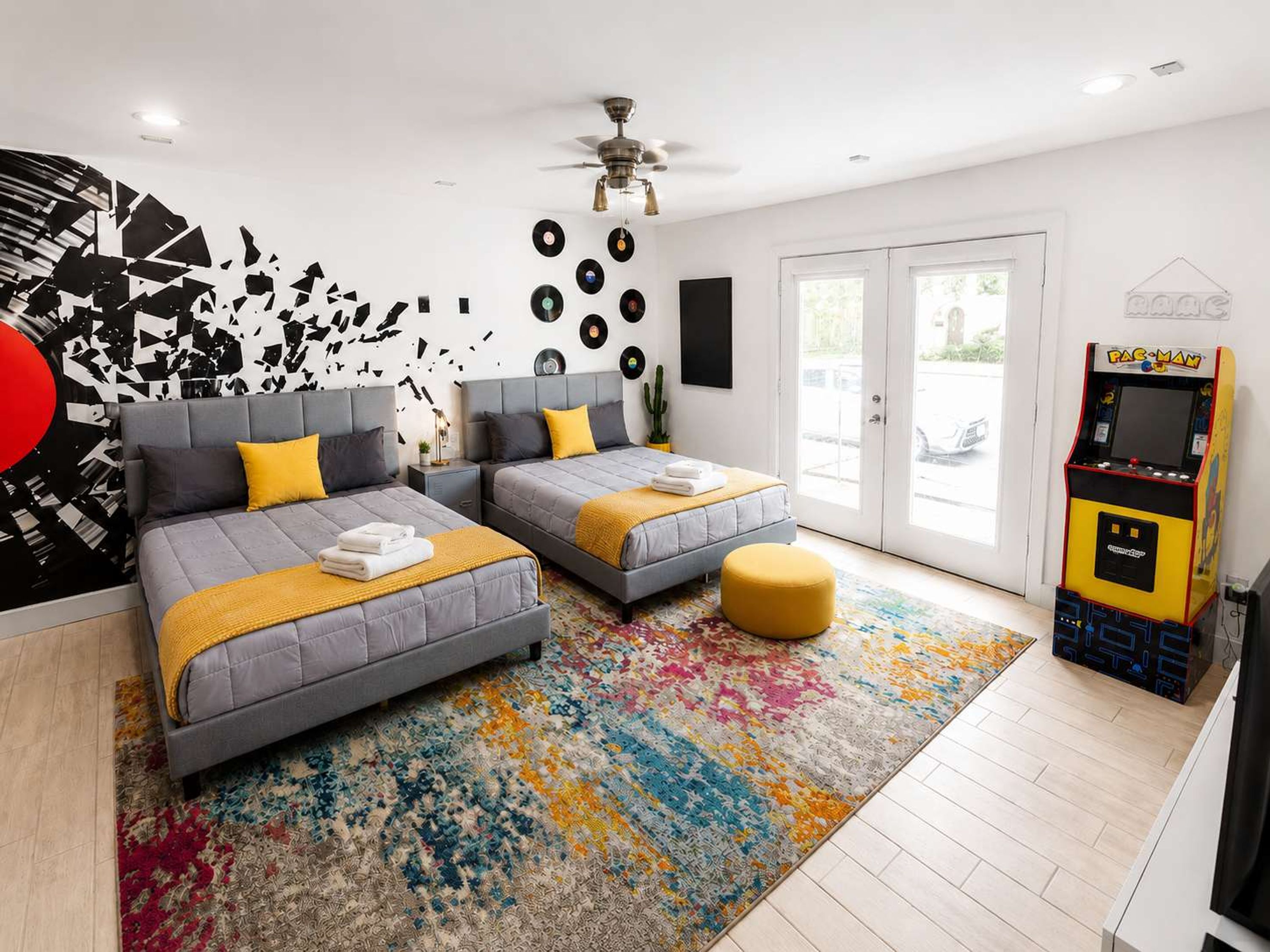 Une chambre moderne avec deux lits, un ventilateur de plafond, une borne d’arcade Pac-Man et une grande fenêtre.