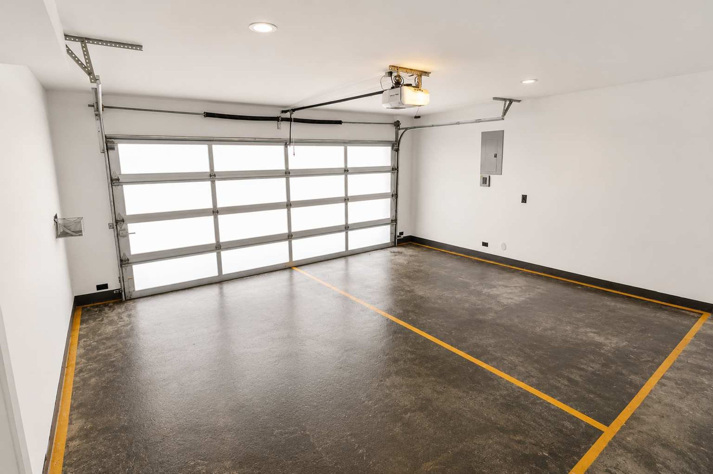 Eine saubere, leere Garage mit einer großen Garagentür, einem Betonboden mit gelben Markierungen und Deckenleuchten.