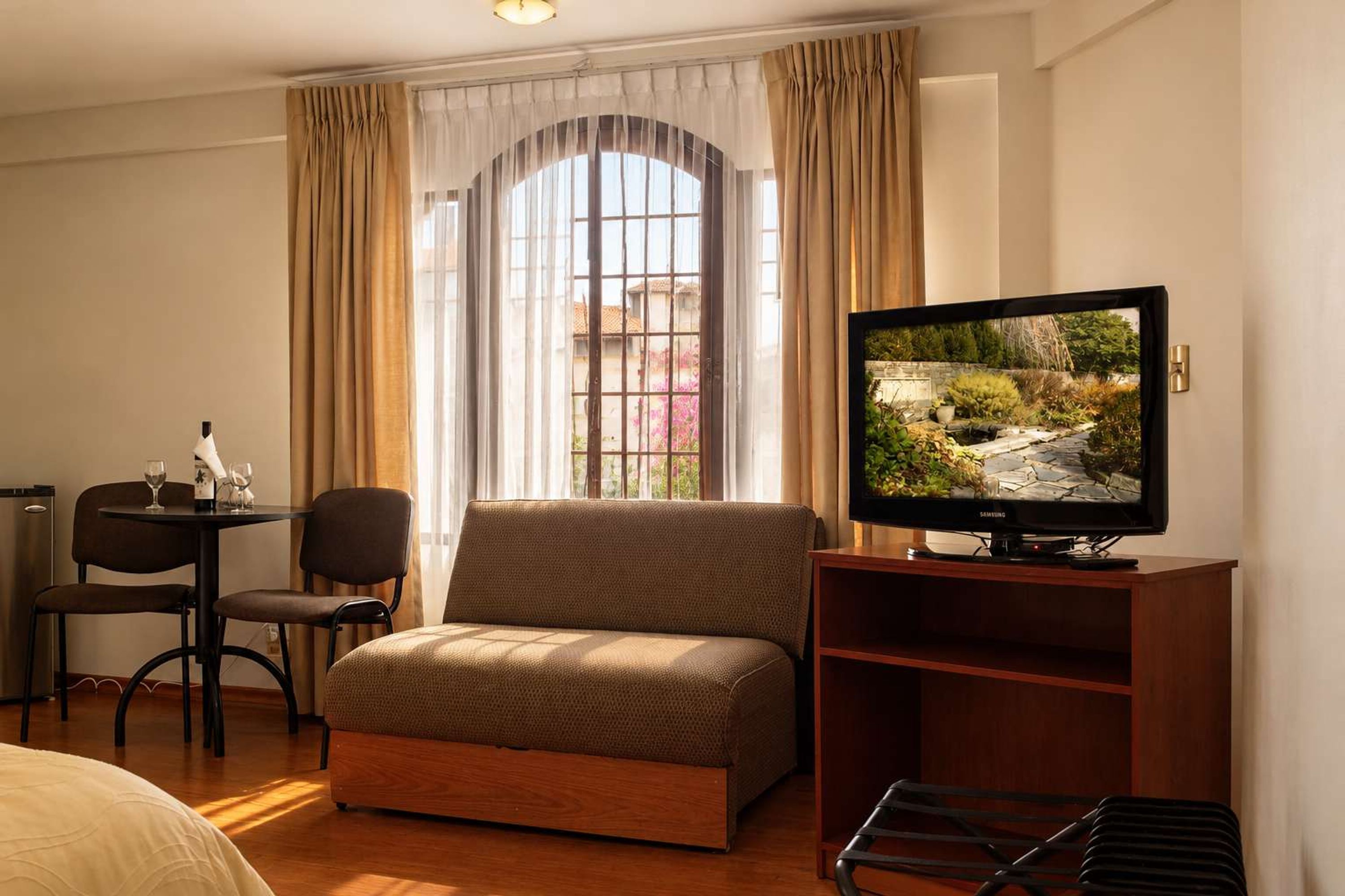 Habitación de hotel con sofá, televisor sobre un mueble de madera, una mesa de comedor con sillas y una ventana con cortinas.
