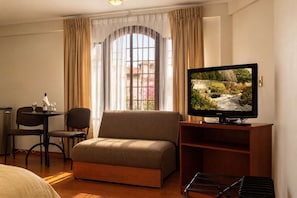 Habitación de hotel con sofá, televisor sobre un mueble de madera, una mesa de comedor con sillas y una ventana con cortinas.