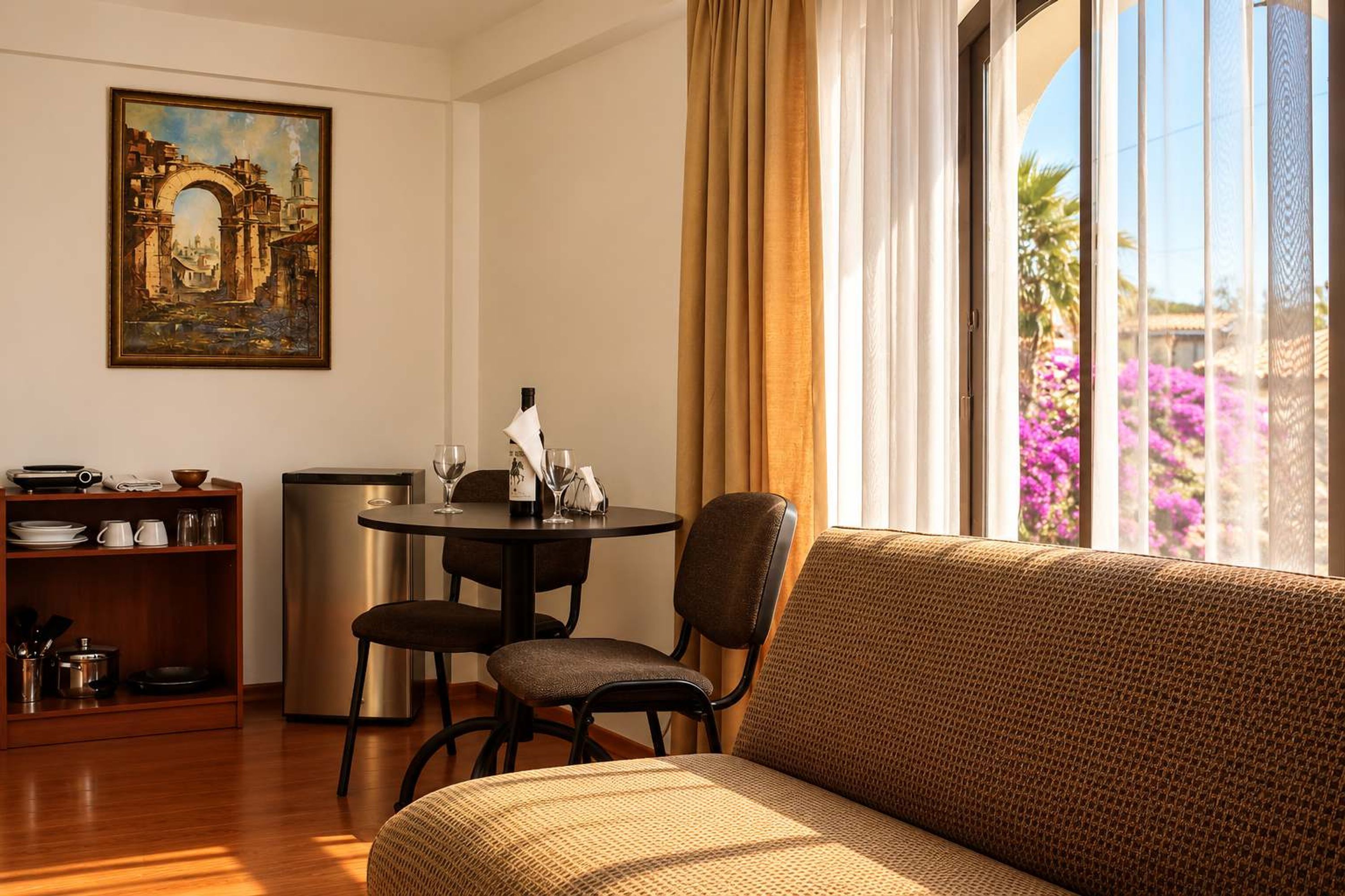 Habitación de hotel con sofá, mesa de comedor, sillas, un cuadro y vista a flores a través de la ventana.