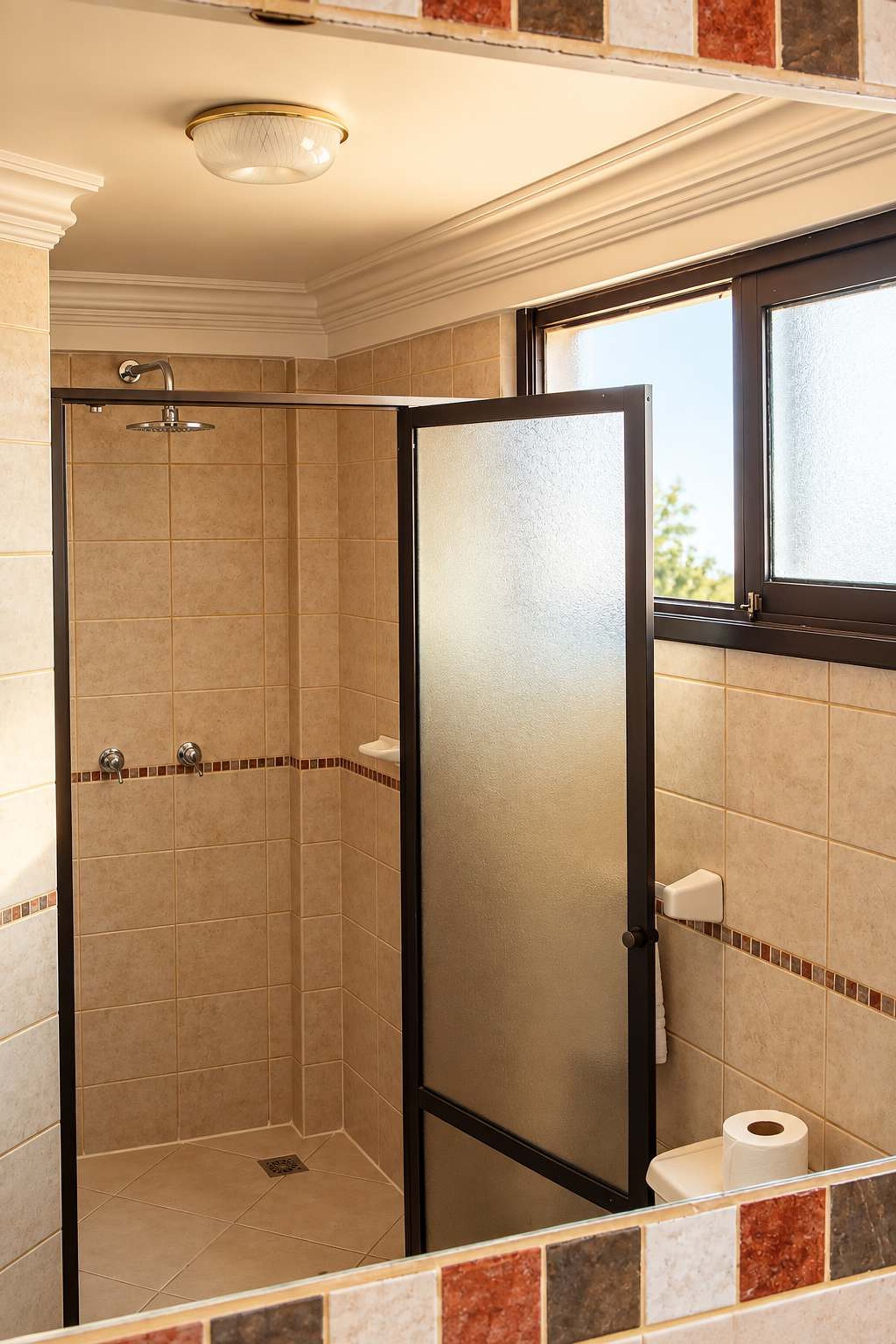 A shower with a glass door, a toilet with a roll of toilet paper, and a window above the toilet.