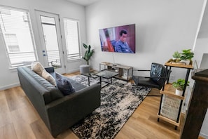 Una sala de estar moderna con un sofá, una mesa de centro y un televisor. Hay ventanas con persianas, una planta y una pequeña mesa con objetos encima.