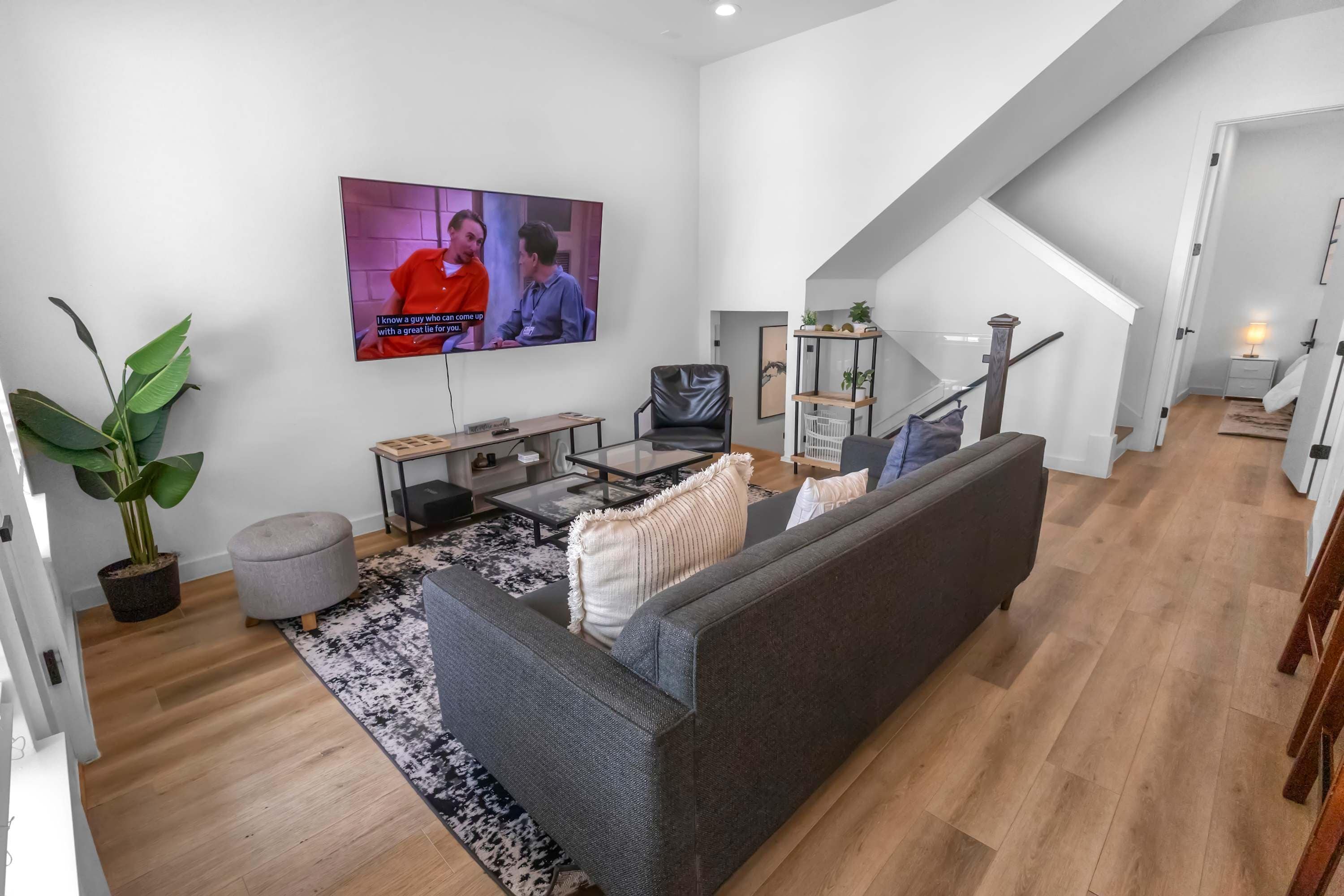 A modern living room with a grey sofa, a coffee table, a TV mounted on the wall, and a staircase leading to an upper level.