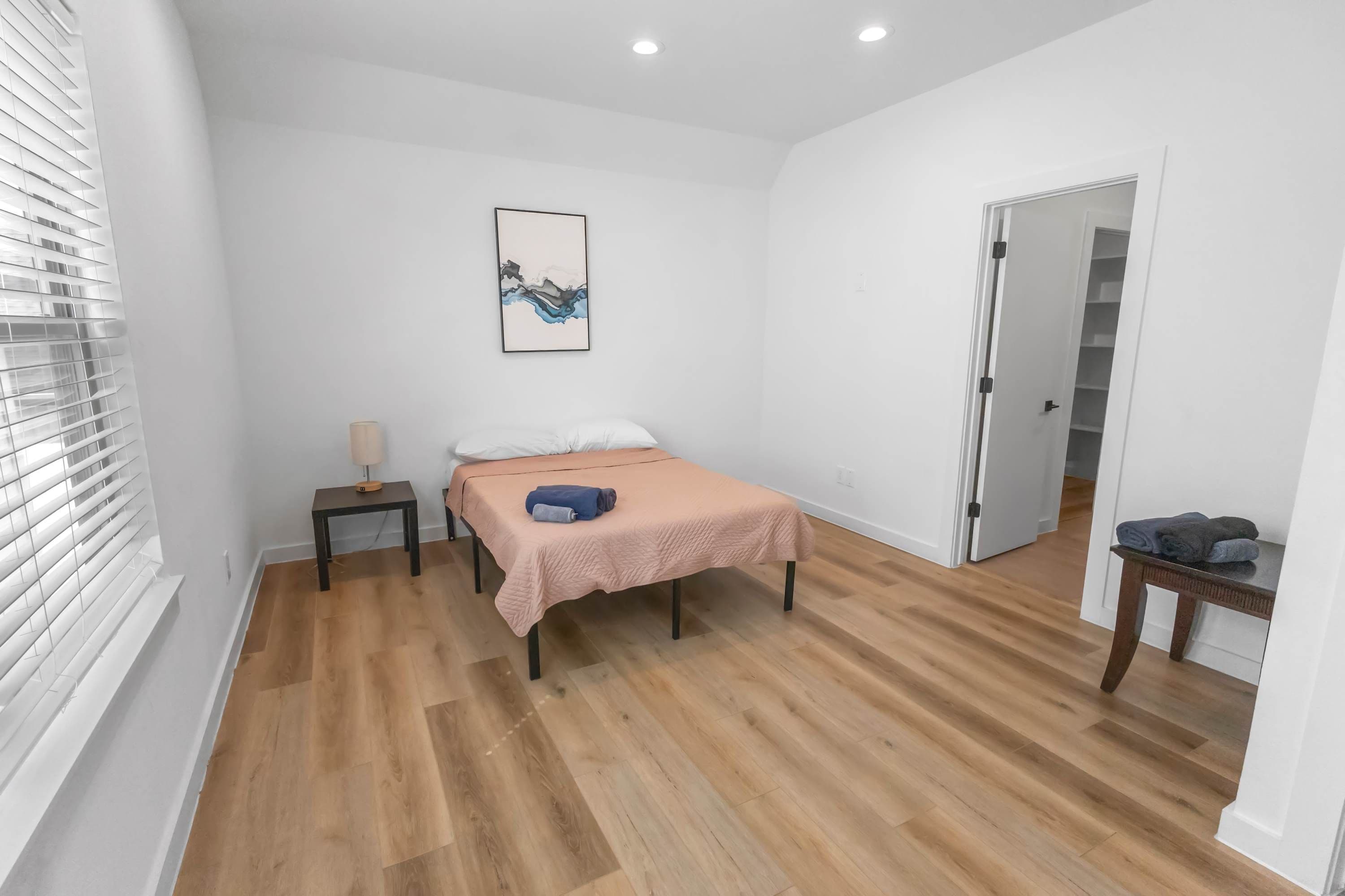 A bedroom with a bed, a bedside table, a lamp, a door, and a hallway.