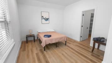 A bedroom with a bed, a bedside table, a lamp, a door, and a hallway.